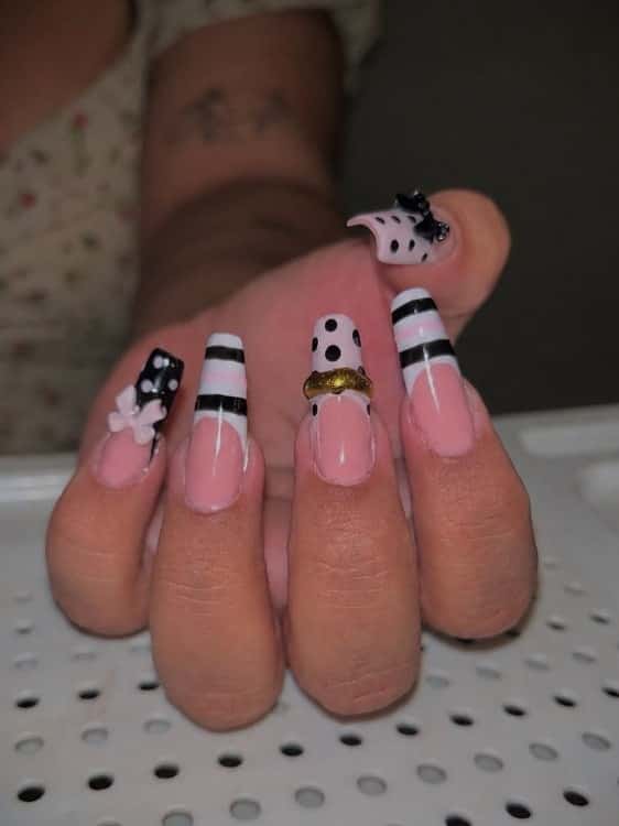 Mão com unhas longas quadradas em base rosada, francesinha listrada em preto e branco, bolinhas, um laço em relevo e um piercing dourado